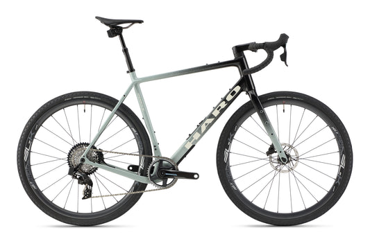 The Buzzard Carbon Top Shelf from Haro Bikes is a sleek, modern road bike with a light gray and black carbon frame. It features drop handlebars, large black wheels with disc brakes, a SRAM Force-like gear system, and gravel tires for versatile terrains. The brand name is prominently displayed on the frame.