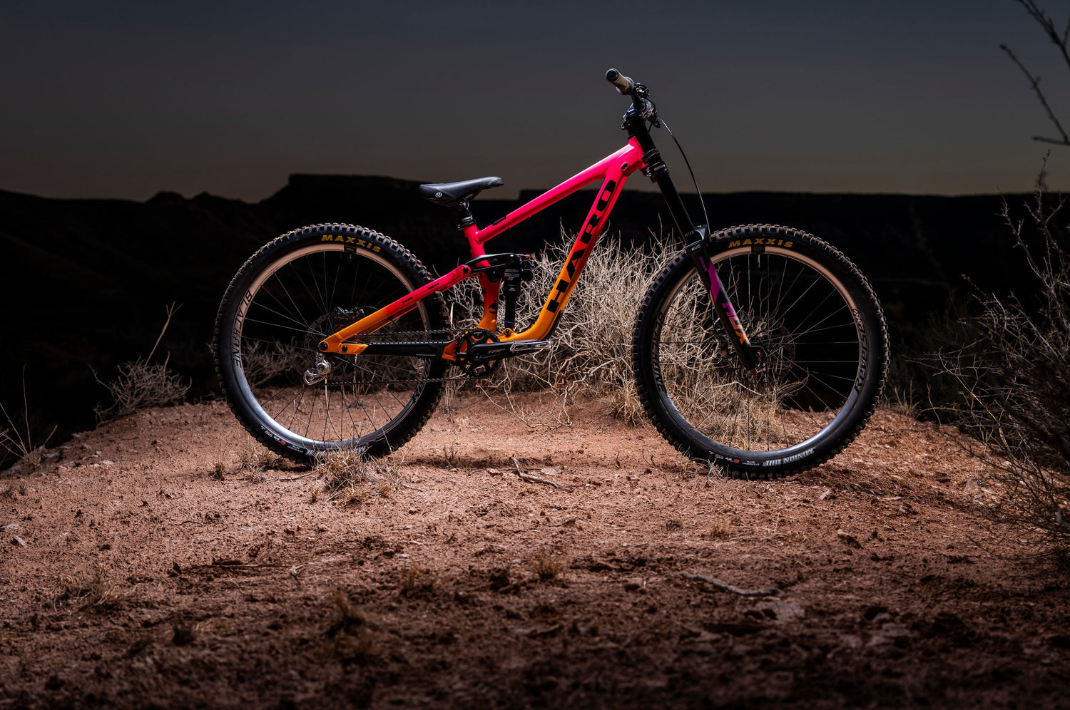 A brightly colored orange and pink mountain bike stands on a dirt mound at dusk, with rugged terrain and a darkening sky in the background.