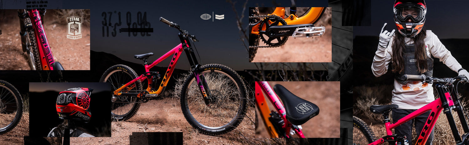 A brightly colored pink and orange mountain bike is parked on a dirt patch with desert terrain and mesas in the background at dusk.