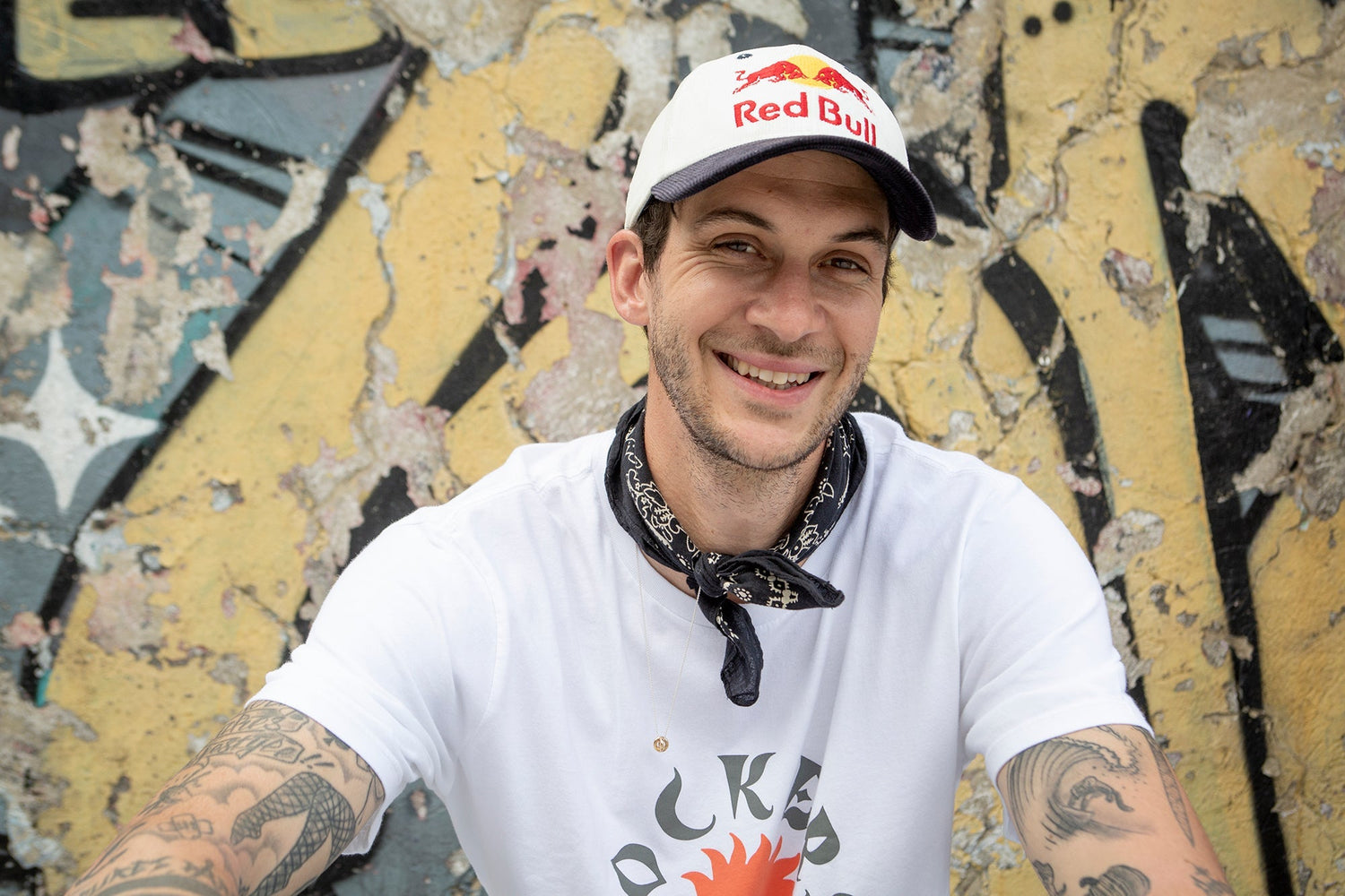 A smiling man wearing a Red Bull cap, white t-shirt, and black bandana sits in front of a weathered graffiti wall. His arms show various tattoos.