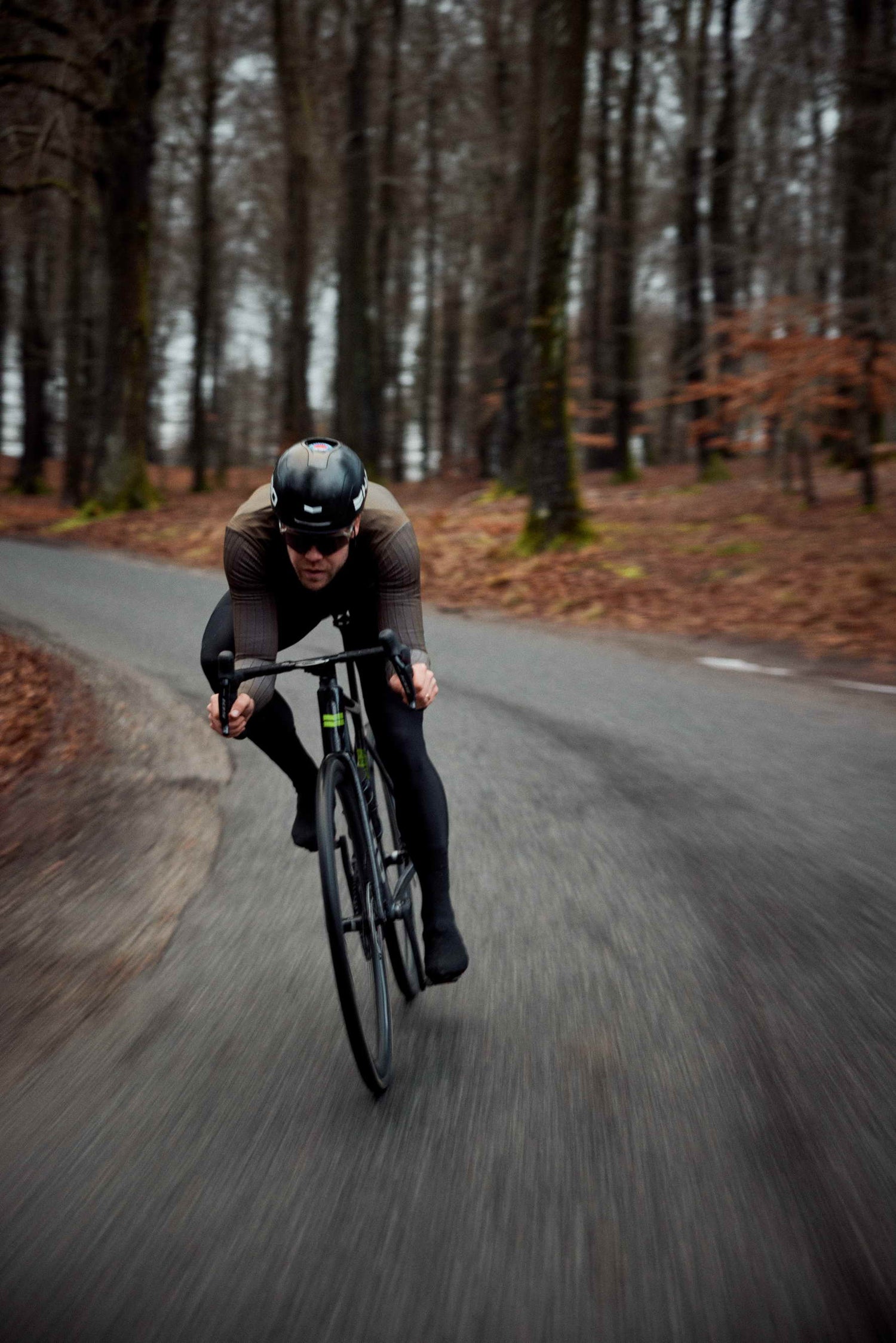 A cyclist wearing a helmet and sunglasses rides quickly around a bend on a forest road, surrounded by leafless trees and brown foliage, with motion blur emphasizing speed.