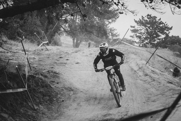 A mountain biker in teal gear rides along a steep, curved wooden wall on a dirt trail, with grassy terrain in the foreground and misty mountains in the background.
