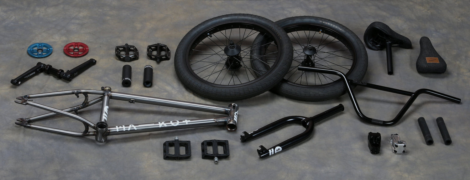 Disassembled BMX bike parts laid out on a gray surface, including a silver frame, wheels, handlebars, seat, cranks, pedals, and various other components arranged neatly.