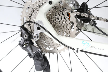 Close-up of the rear derailleur and cassette showcasing the Trail-Geometrie design on a Double Peak Comp 27.5 by Haro Bikes. The white frame contrasts with the black derailleur, connected by a cable, highlighting the precision mechanics of its gear system.