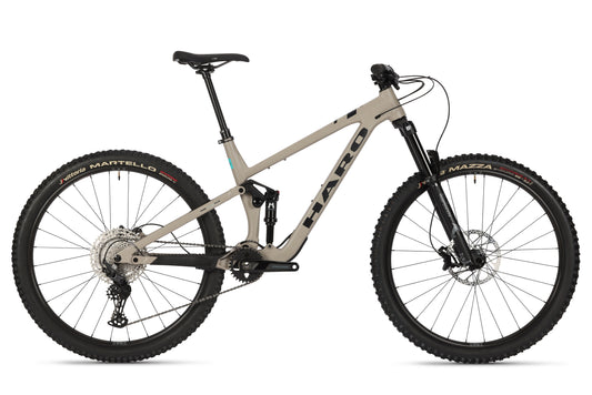 The Daley Alloy 2 from Haro Bikes is a beige All-Mountain-29er featuring a robust aluminum frame, front and rear suspension, 29-inch wheels, disc brakes, and black saddle and handlebars. The tires feature red text reading Martello and Mazza.