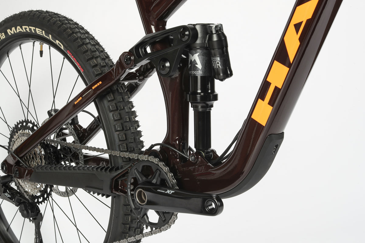 Close-up of the Greer Alloy 1s rear suspension, chain, and crankset featuring a progressive Federung. The Haro Bikes frame flaunts a sleek dark finish with orange branding, set against a plain white background highlighting the tire tread.