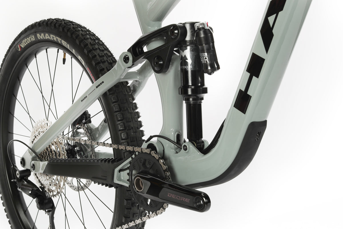 Close-up of a gray Haro Bikes Greer Alloy 2 mountain bikes drivetrain and rear suspension system, featuring a Deore crankset, chain, and rear shock beneath the frame. Its back tire boasts textured tread for off-road adventures.