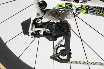 Close-up of a Haro Bikes Rivette Carbon LTDs rear derailleur and cassette, connected by a metallic chain to the gear system. Against a white background highlighting part of the rear wheels spokes, the limited edition frame features white with green lettering designed by Mark Ward.