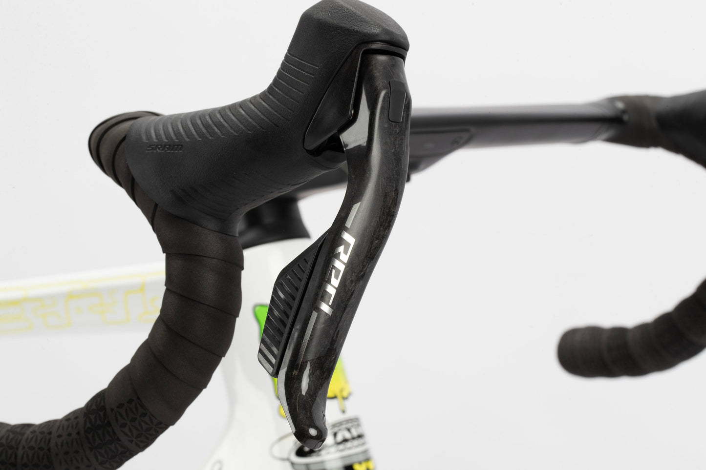 Close-up of a Haro Bikes Rivette Carbon LTD handlebar with black grip tape and a SRAM gear lever. The white frame, designed by Mark Ward, features a partial design with yellow and green accents, standing out against the plain white background.