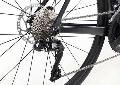 Close-up of the Rivette Carbon 2s rear derailleur and cassette. The silver cassette features multiple cogs, while the sleek black derailleur by Haro Bikes is frame-mounted with internal cable routing. The white background enhances the detailed mechanics and chain links.