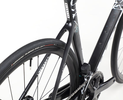 Close-up of a sleek black Rivette Carbon 2 by Haro Bikes, focused on the rear wheel and frame. The tire reads Vittoria Rubino Pro G 2.0, with high-pitch carbon fiber showcasing a unique pattern. The chainring and part of the drivetrain are visible.