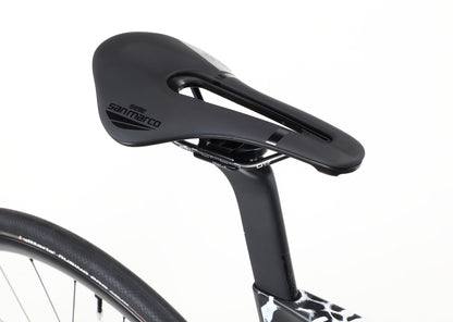 Close-up of a black Selle San Marco saddle on the Rivette Carbon 2 by Haro Bikes. The sleek seat post complements the carbon fiber frame with internal cable routing, and the saddles central cutout ensures comfort, with part of the bike wheel and frame visible below.