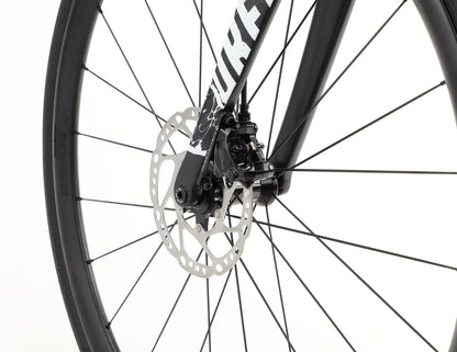 A close-up of the Rivette Carbon 2 race-ready wheel by Haro Bikes showcases a disc brake on the metal rotor, mounted at the hub. The black fork with white lettering stands out against a plain background, featuring internal cable routing and sleek spokes radiating stylishly outward.