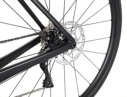 A close-up of the Rivette Carbon 2 by Haro Bikes showcases the rear wheels spokes, disc brake, and rear derailleur. The metal components contrast with the sleek black frame and carbon fiber, highlighting this road bikes mechanical details.