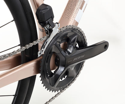 Close-up of the Haro Bikes Rivette Carbon 2s drivetrain, showcasing a black chainring and 105 crankset. The metallic rose gold frame features sleek internal cable routing, with part of the rear wheel and tire visible.