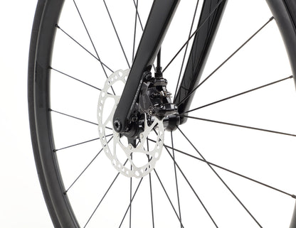 Close-up of the Rivette Carbon 2 by Haro Bikes, featuring a black bicycle wheel with disc brake mechanism on a race-ready road bike. Visible spokes and tire stand against a white background, highlighting its sleek design and internal cable routing.