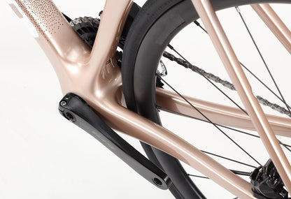 Close-up of the Rivette Carbon 2 by Haro Bikes, featuring a metallic beige frame with crank arm, rear wheel, and chain in view against a white background. The bike showcases internal cable routing and a sleek, modern race-ready road design.