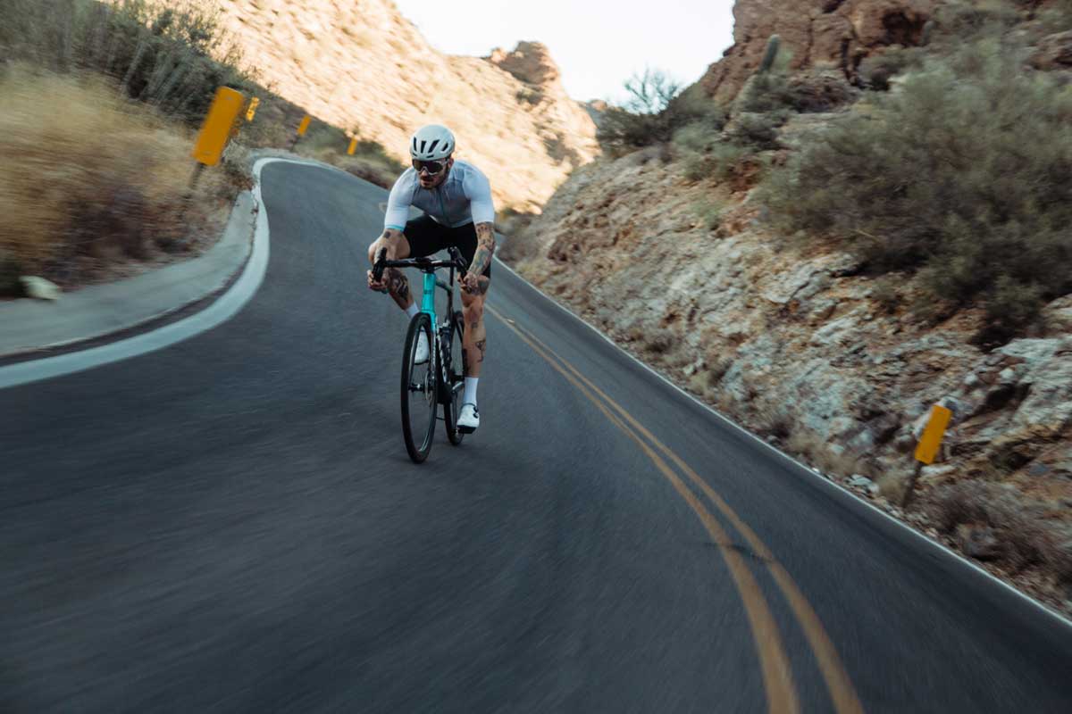 A cyclist in a helmet and sunglasses rides a road bike down a winding mountain road with rocky terrain and sparse vegetation under a clear, sunny sky