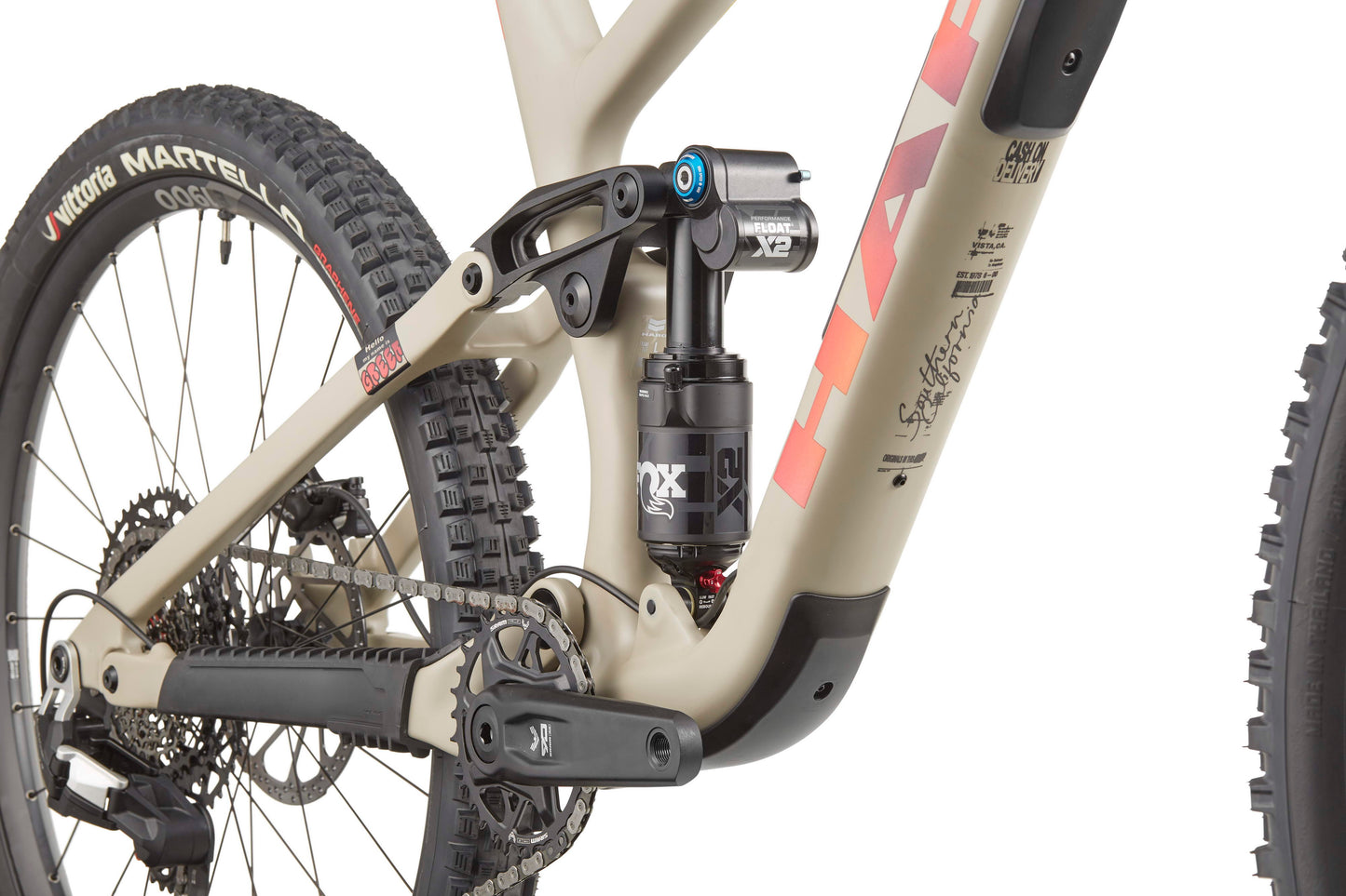 Close-up of Haro Bikes Greer Carbon 3’s rear suspension, drivetrain, and wheel on a beige frame with red graphics for trail agility.
