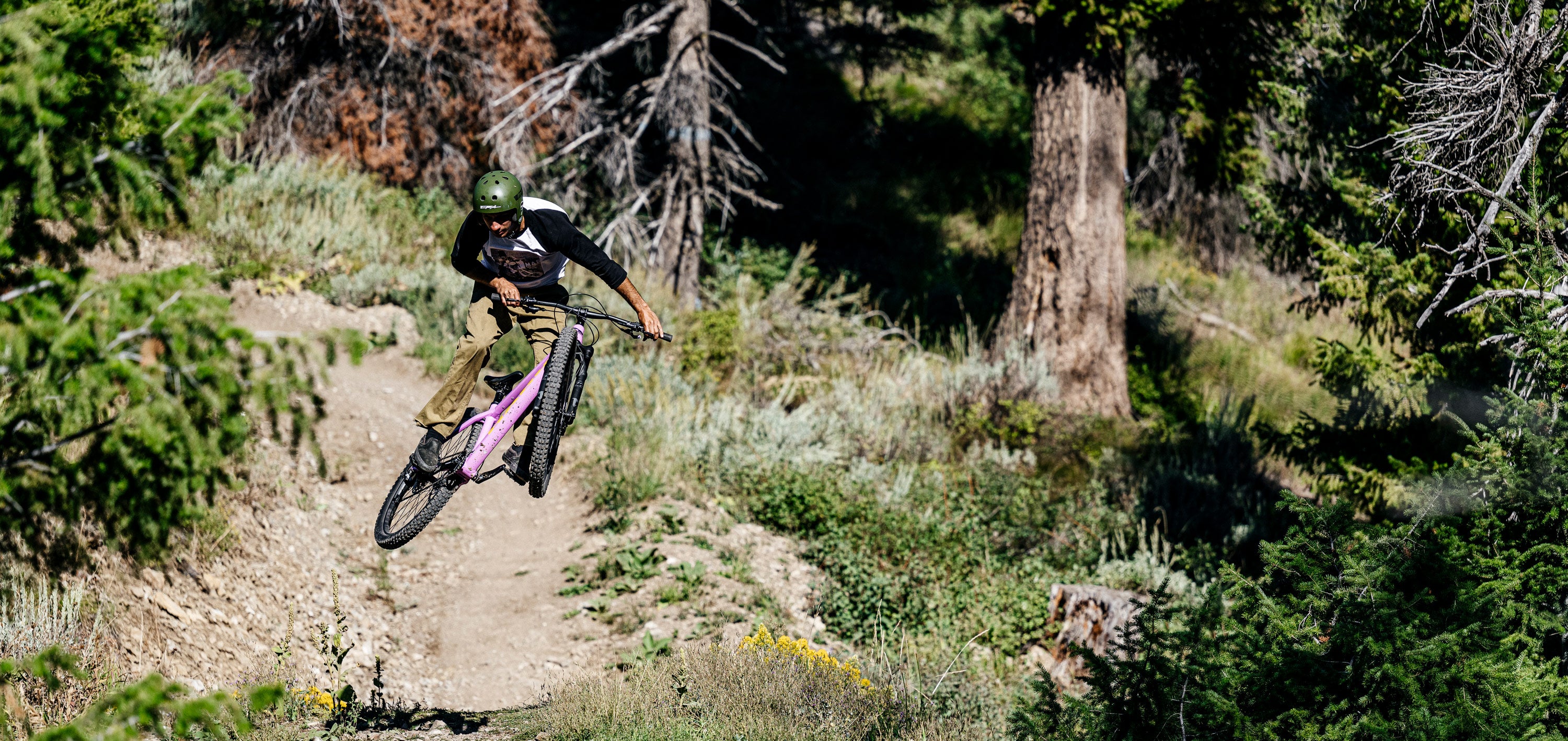 A person wearing a helmet is performing a jump on a pink mountain bike along a forest trail. They are surrounded by tall trees and greenery under sunlight.