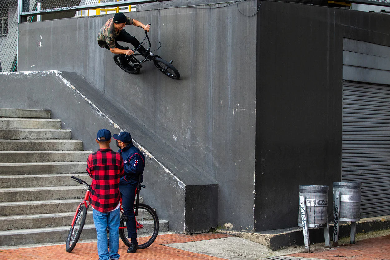 A BMX rider performs a wall ride on a tall concrete structure while two people watch from below, one in a red checkered shirt and cap, near stairs.