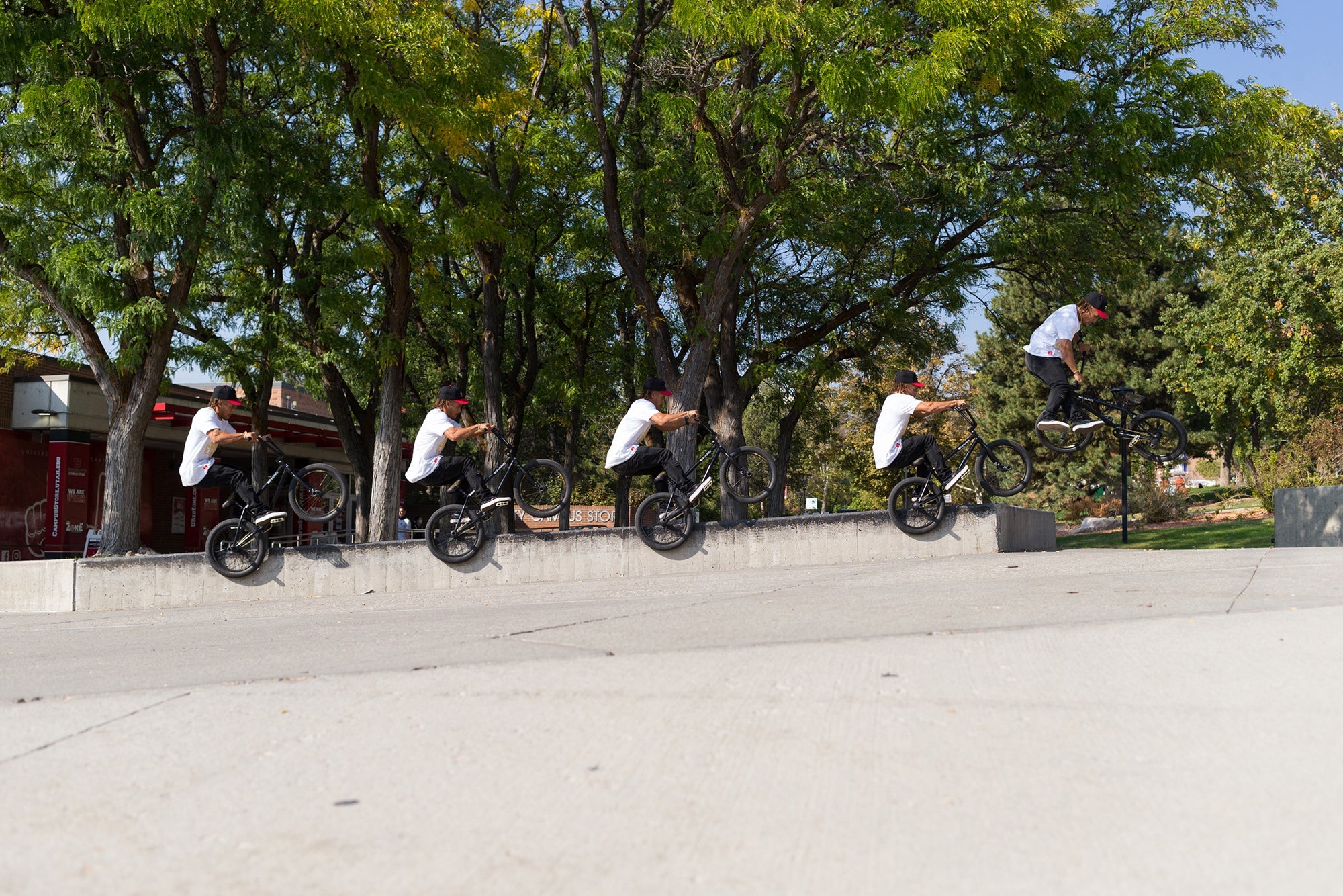 A series of five images shows a person performing a BMX trick, jumping off a concrete ledge in a park. The images capture different stages of the jump against a backdrop of green trees and a clear sky.