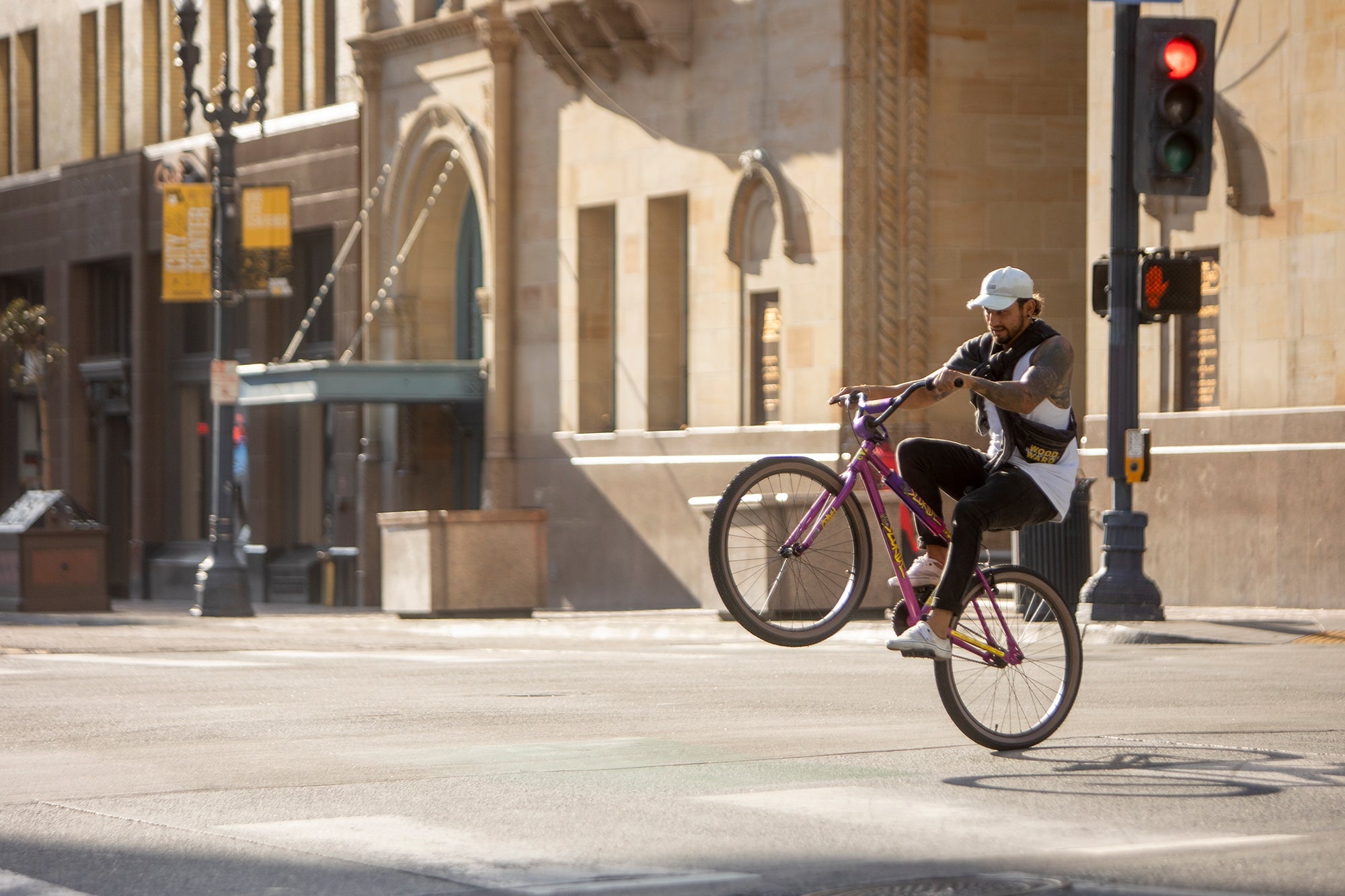 A person performs a wheelie on a pink bicycle while crossing an intersection in a city. They are wearing casual clothes and a helmet. The background features a tall building, traffic light, and street lamp.