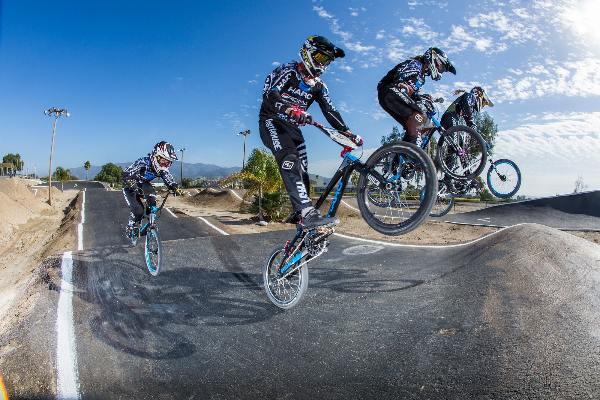 Four BMX riders in black and white uniforms perform jumps on a dirt track under a clear blue sky. The scene captures action and speed, with a backdrop of trees and distant mountains.