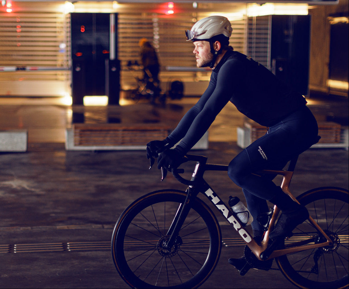 A cyclist wearing a helmet and dark athletic gear rides a road bike in a dimly lit underground parking area. A person stands in the background near a stroller. Warm lights illuminate the space.