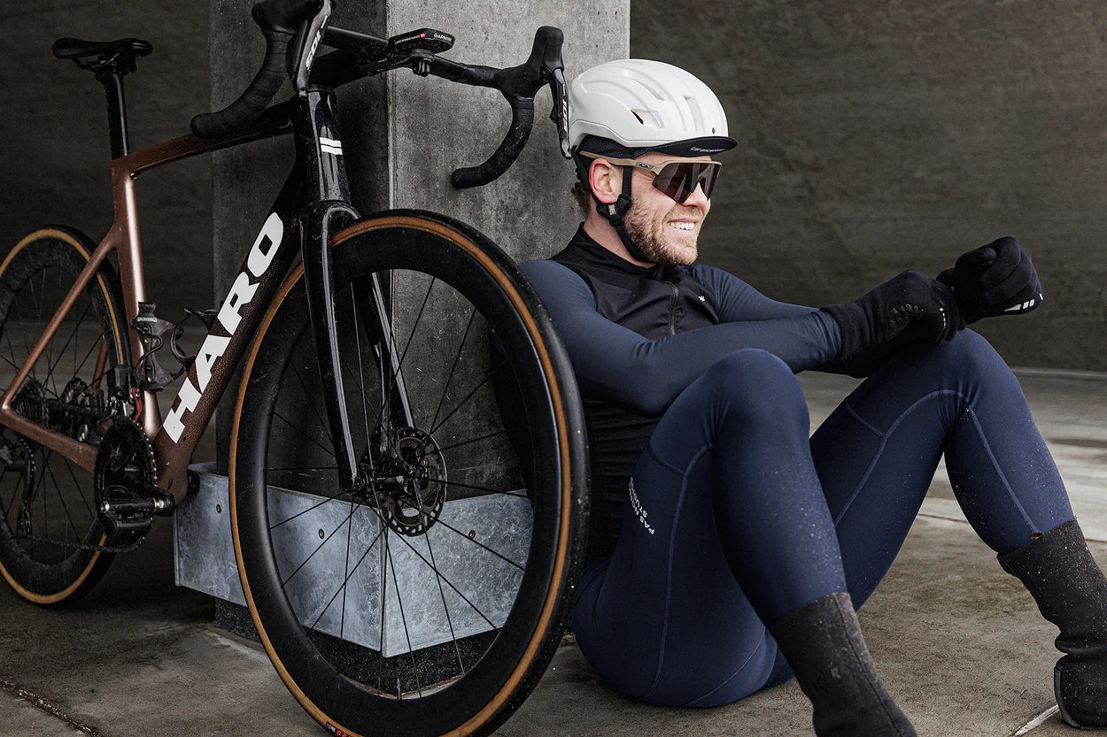 A cyclist wearing a white helmet, sunglasses, and dark cycling attire sits on the ground resting against a concrete pillar, smiling. A brown bicycle is leaning against the pillar beside him.