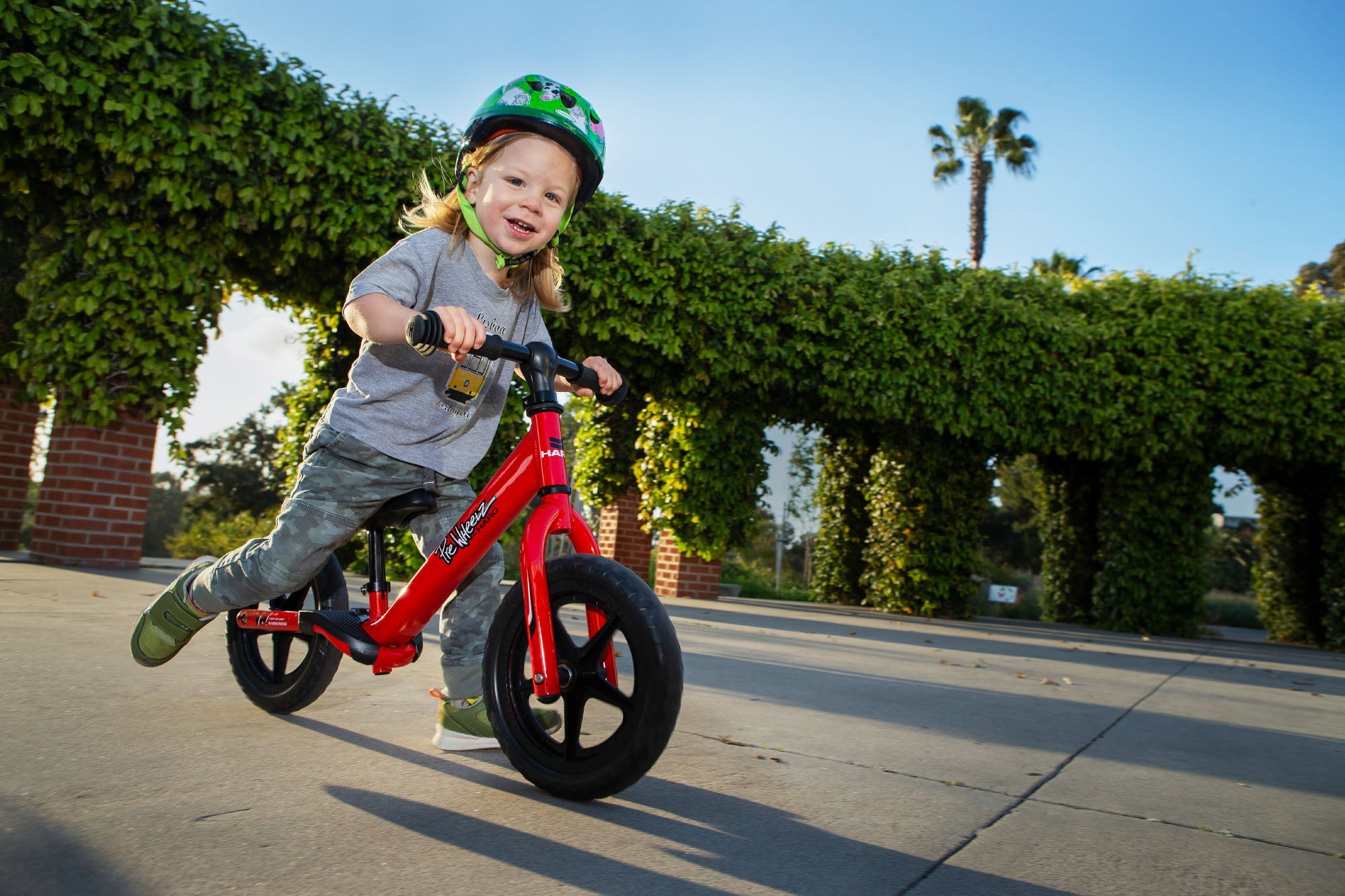 A young child wearing a green helmet is happily riding a red balance bike on a sunny day. In the background, there is a green vine-covered arbor and a clear blue sky. The child is dressed casually in a gray shirt and camo pants.