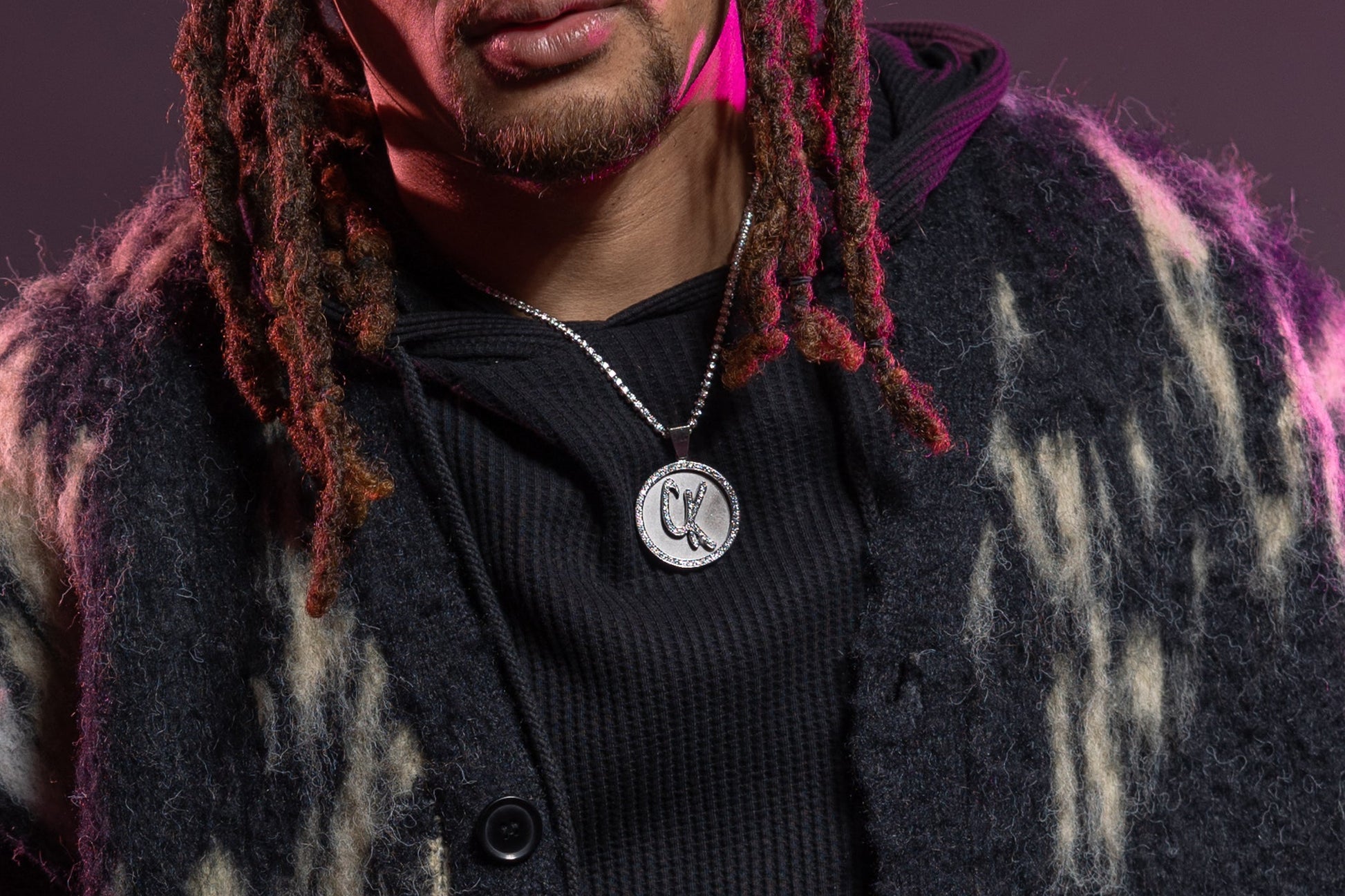 A person with dreadlocks wears a textured black coat, a round pendant, and holds a Premium CK Top Load Stem V3.