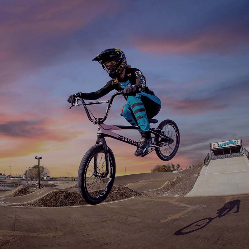 A person wearing a helmet and protective gear performs a jump on a BMX bike at a track with mounds of dirt. The sky is colorful with shades of pink and purple as the sun sets in the background.