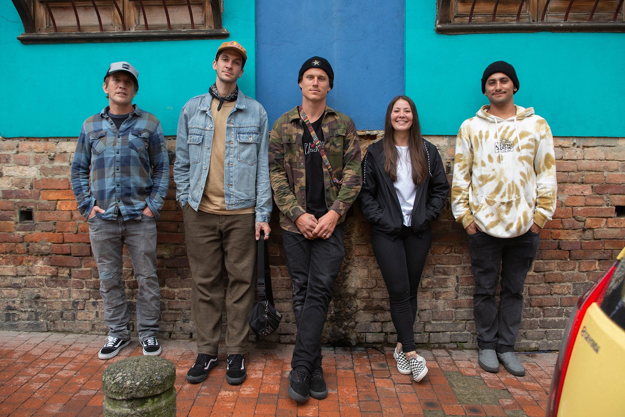 A group of five people stands against a brick and vibrant blue wall. They are casually dressed, one in a plaid shirt, another in denim, with others in jackets and hoodies. They appear relaxed and are standing on a brick sidewalk.