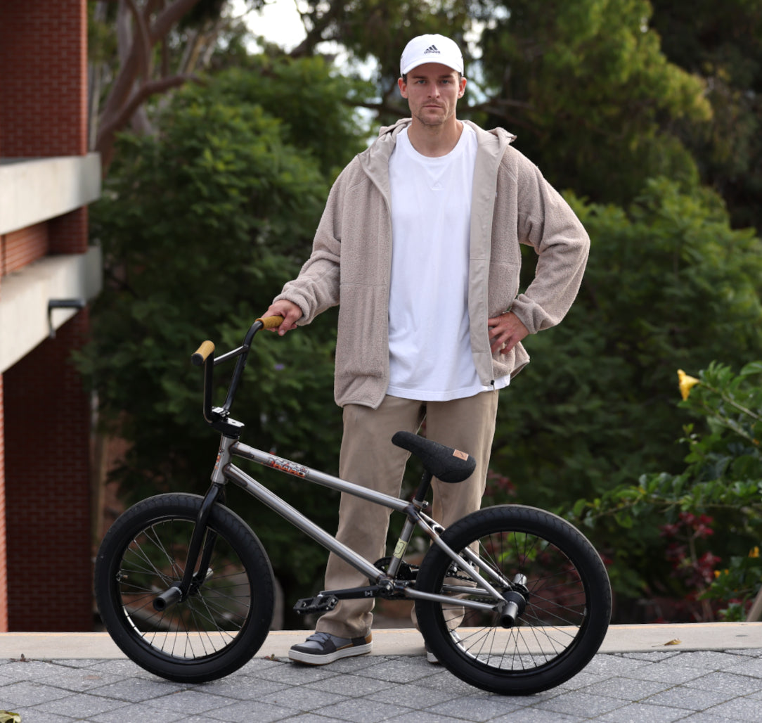 A person in a beige jacket and white cap stands outdoors next to a BMX bike. Theyre on a paved surface with greenery and a brick building in the background.