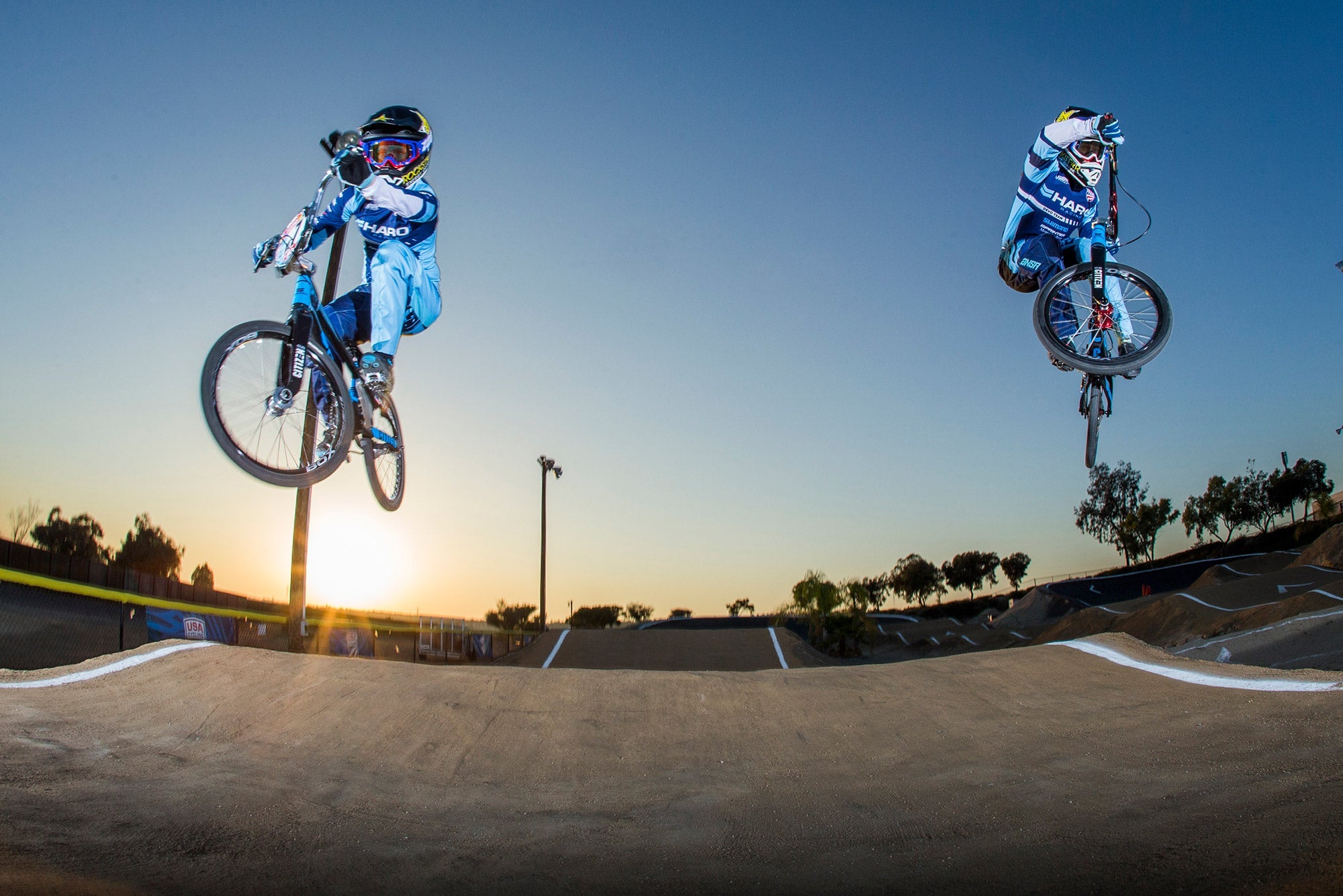 Two bikers in blue uniforms perform jumps on a BMX track at sunset. One rider is mid-air on the left, while the other is airborne on the right against a clear sky. The track is surrounded by trees and a low barrier in the background.