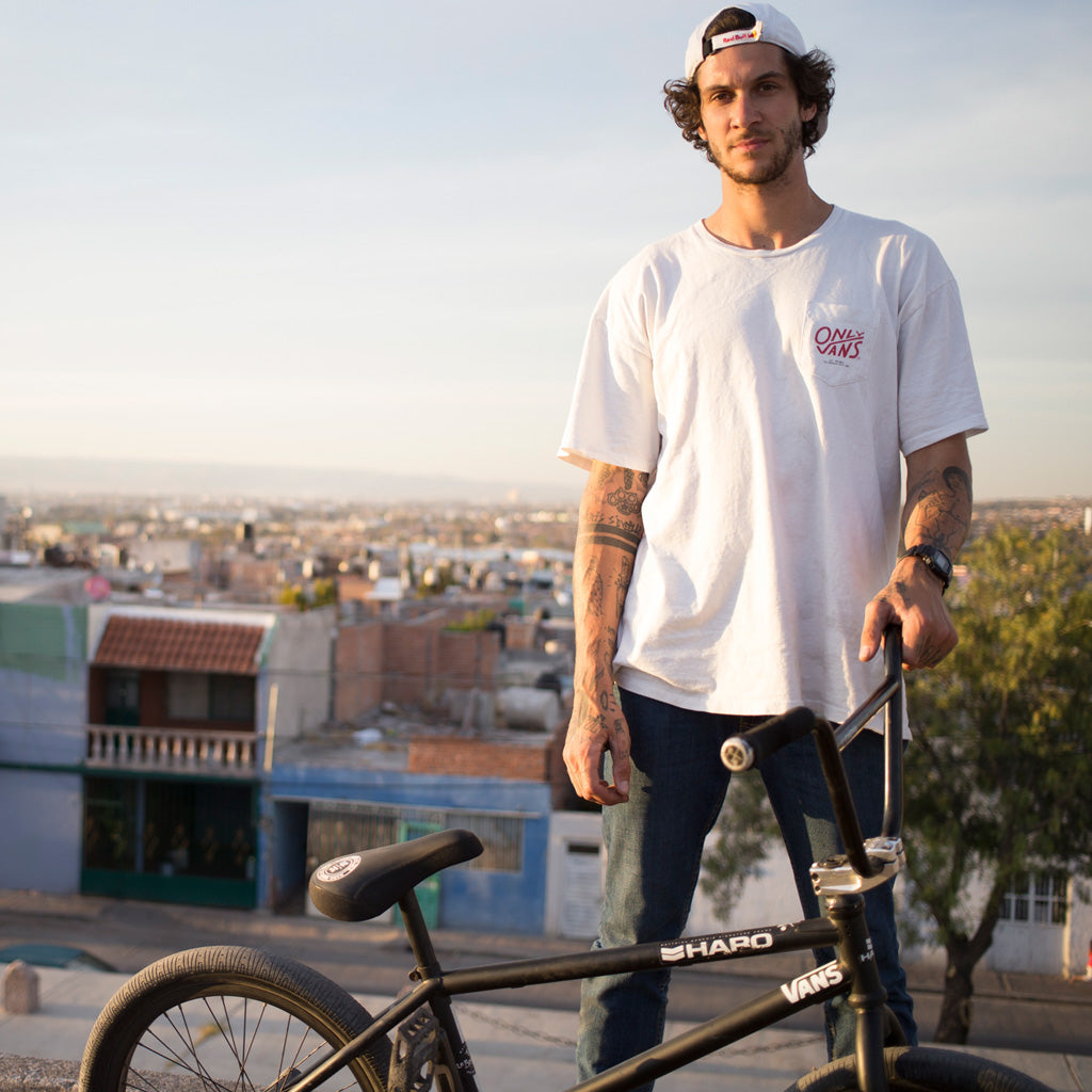 A man wearing a white T-shirt and cap stands next to a black BMX bike, overlooking a cityscape at sunset. He has tattoos on his arms and is holding the handlebar of the bike, with houses and trees in the background.